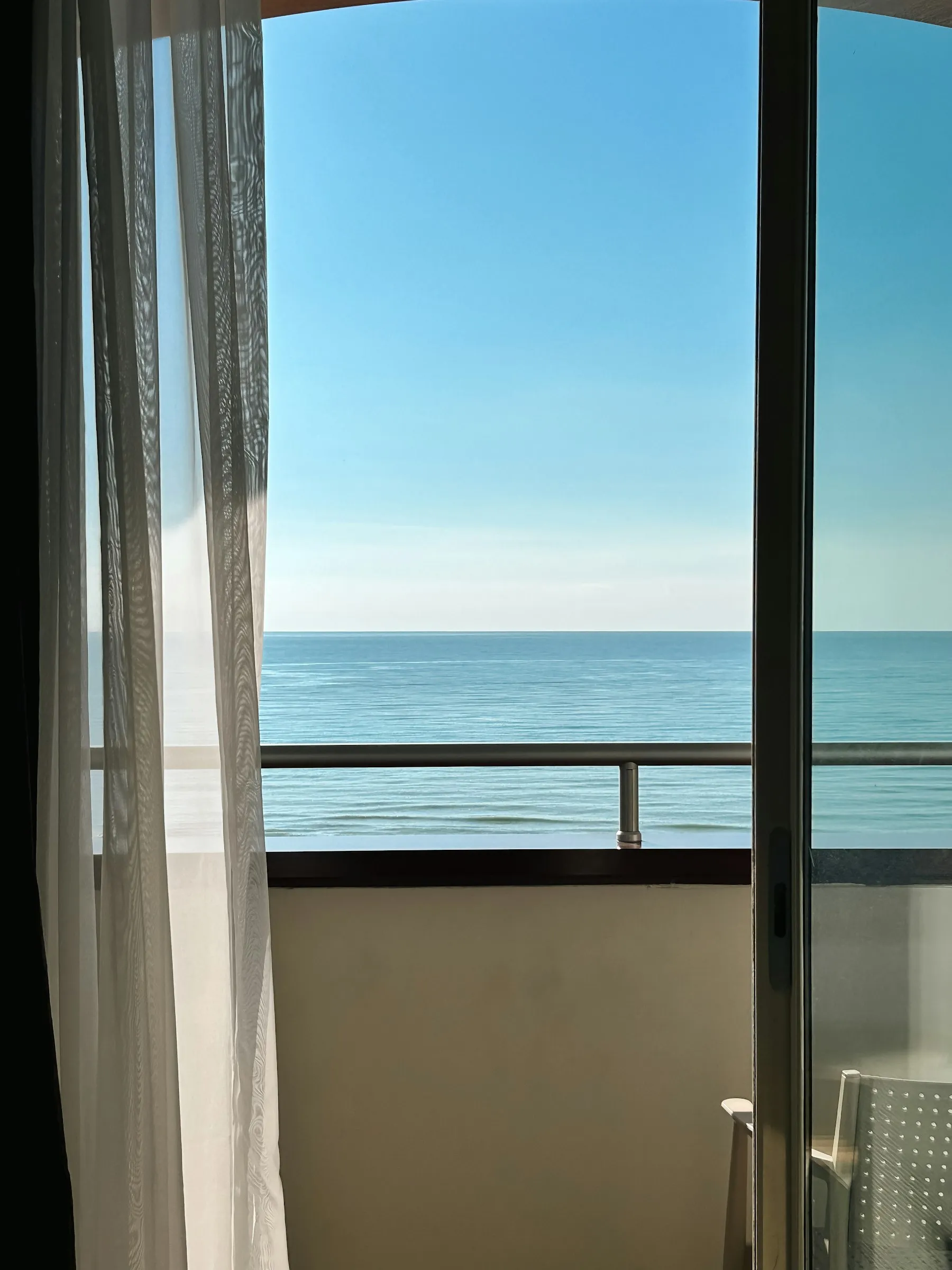 Vista do mar azul pela porta de vidro da varanda, com a cortina leve